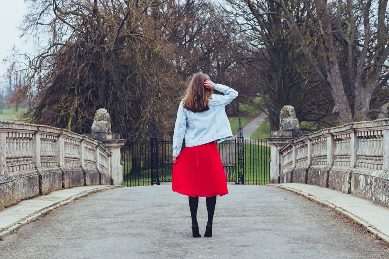 A woman in a red dress and jean jacket stands on a bridge.